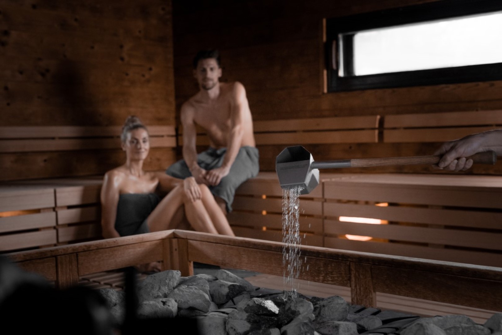 Zwei Personen entspannen in einer holzverkleideten Sauna, während Wasser auf heiße Steine gegossen wird und Dampf aufsteigt.
