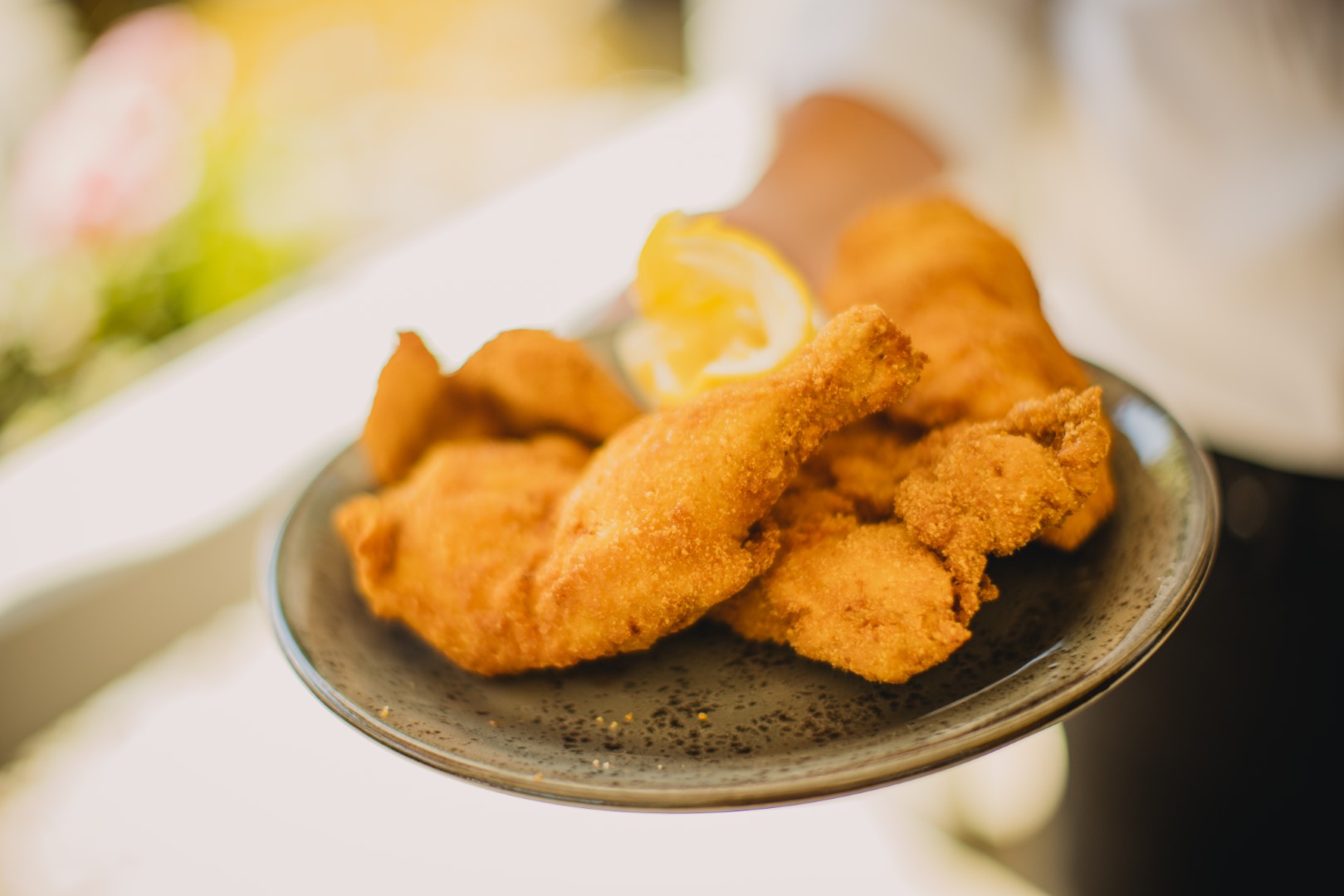 Hand hält einen Teller mit knusprigen, goldbraunen Hühnerschnitzeln, serviert mit einer Zitronenscheibe.