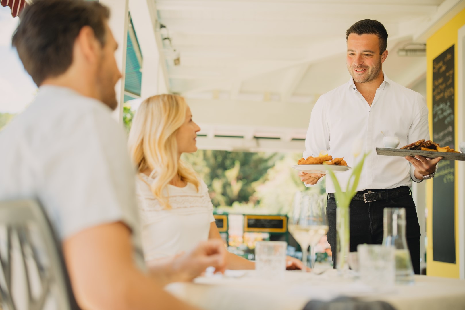 Kellner serviert herzhaftes Essen an ein Paar auf der sonnigen Terrasse des Gasthauses Treiber.