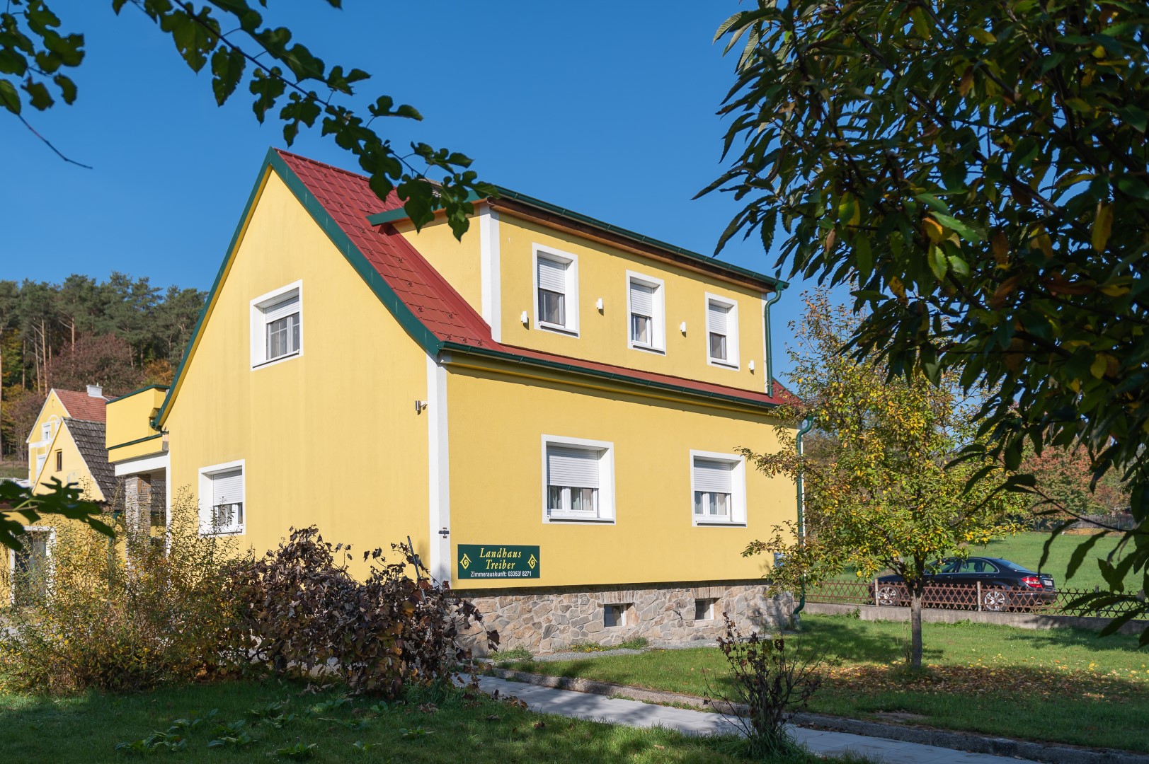 Gelb gestrichenes Landhaus Treiber mit rotem Dach, umgeben von grüner Vegetation und klarem Himmel.