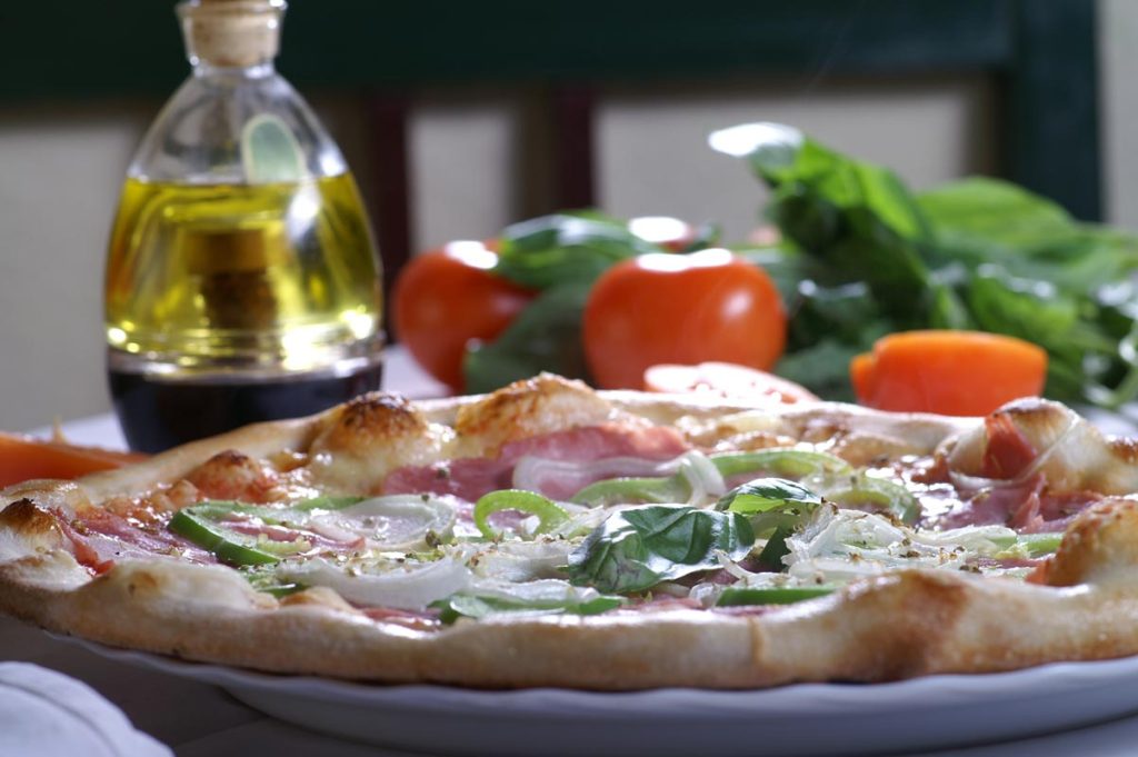 Frisch gebackene Pizza mit Schinken, Paprika und frischen Kräutern, serviert mit Olivenöl und Tomaten im Hintergrund.