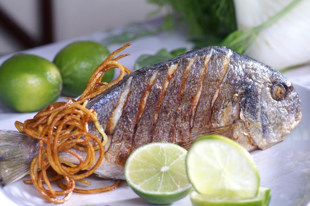 Ein ganzer gegrillter Fisch mit Kräuterfüllung, begleitet von Limettenscheiben und knusprigen Kartoffelspiralen.