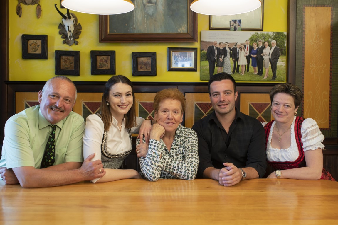 Farbiges Familienportrait von fünf Personen, die in traditioneller und zeitgenössischer Kleidung in einem Speisesaal des Gasthaus Treiber lächeln.