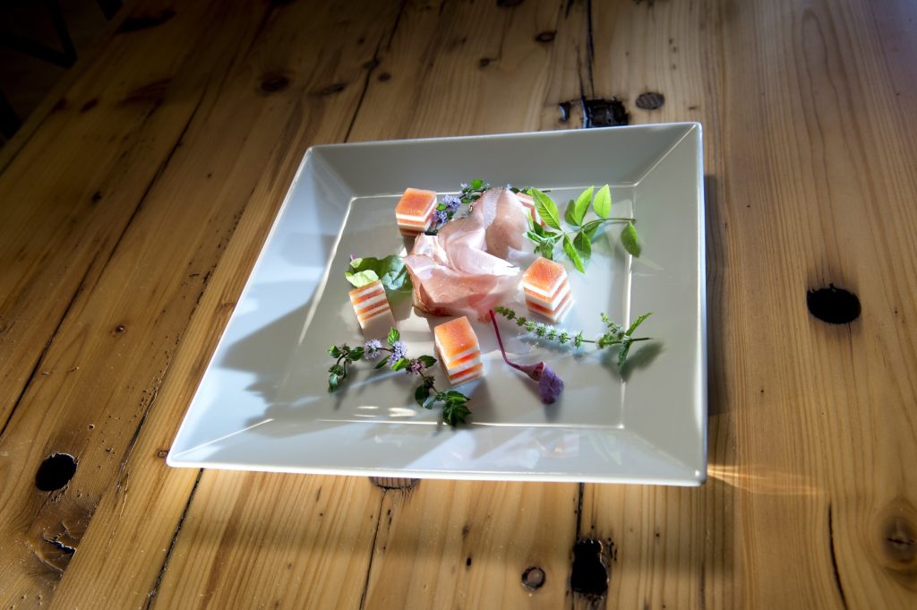 Eine kunstvoll angerichtete Vorspeise mit Prosciutto und Melone auf einem quadratischen weißen Teller, präsentiert auf einem rustikalen Holztisch.
