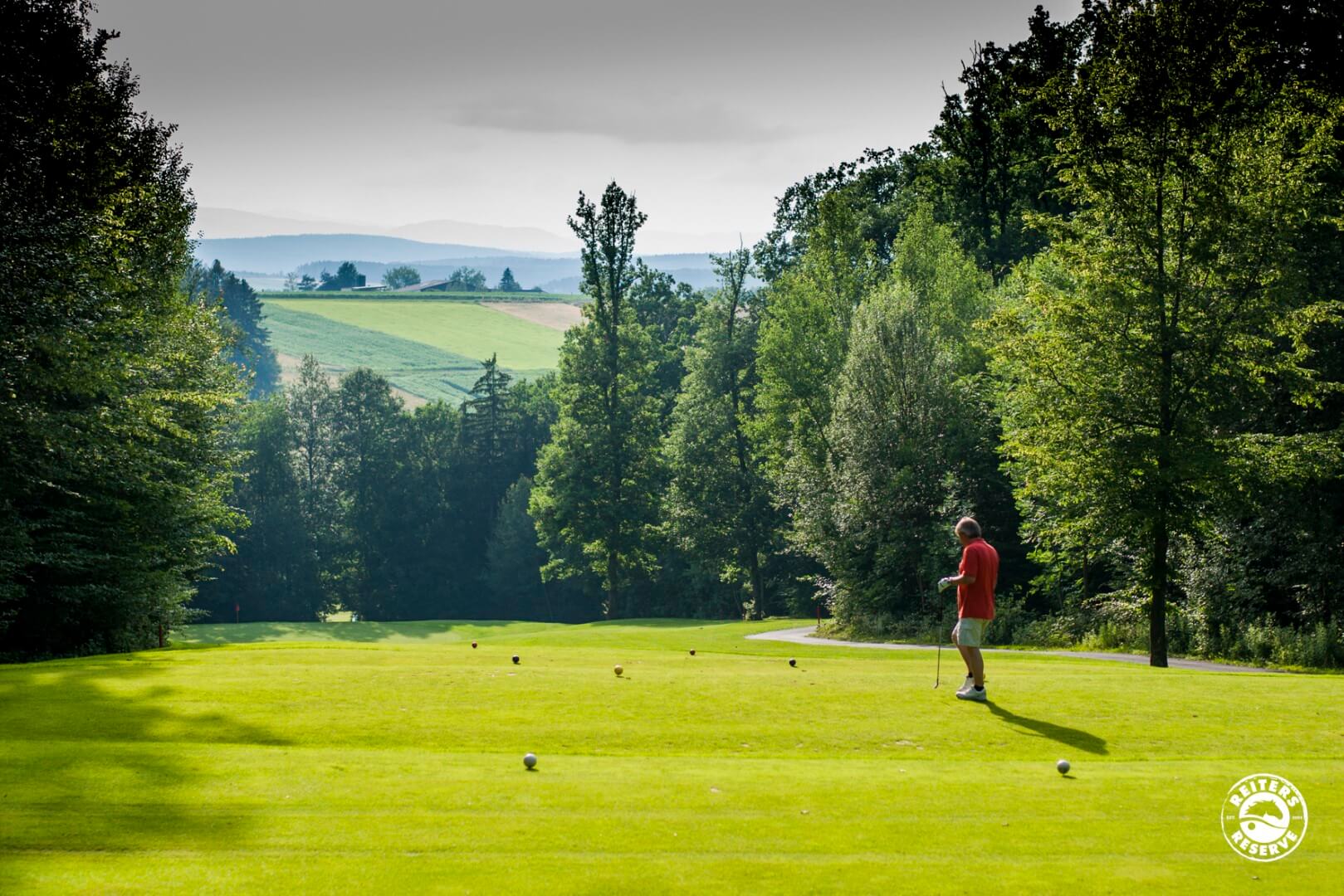 Ein Golfer in rotem Shirt bereitet sich auf dem Abschlagplatz vor, umgeben von dichtem Wald und einer hügeligen Landschaft.