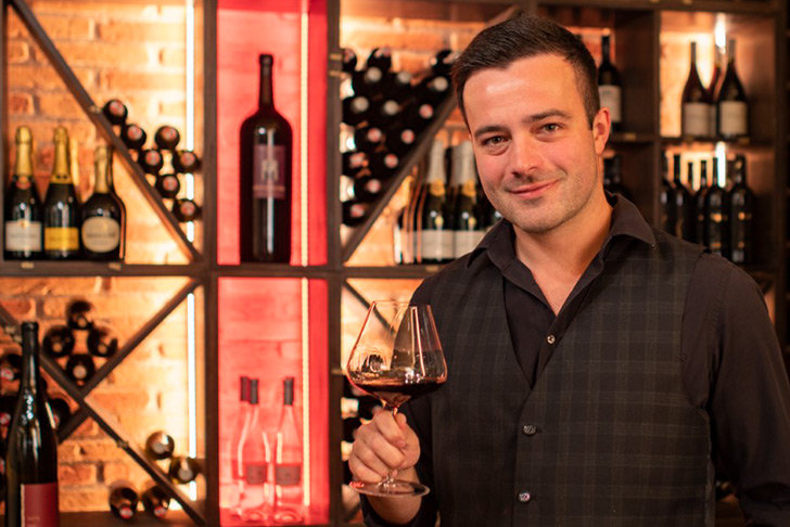 Sommelier Herbert Treiber Jun. im Gasthaus Treiber hält ein Glas Rotwein, umgeben von einer Auswahl an Weinen in der Vinothek.